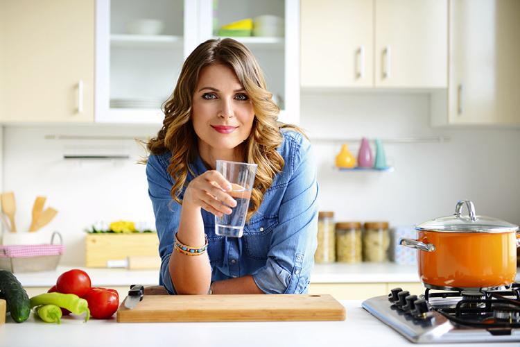 Mulher loira na cozinha com copo de água na mão olhando para a foto