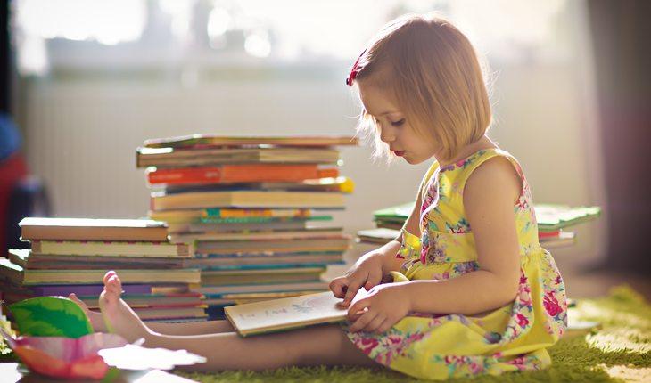criança sentada num tapete verde lendo um livro e vários outros perto dela