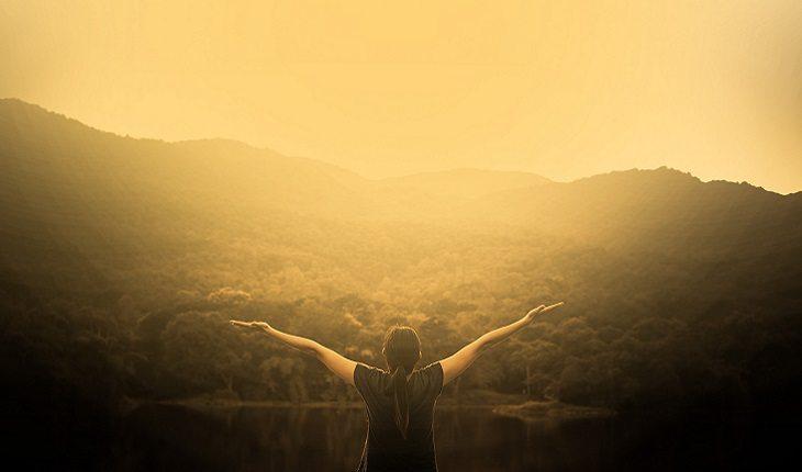 A foto mostra uma mulher com os braços abertos em cima de uma montanha contemplando a paisagem natural, ilustrando uma das leis morais do espiritismo