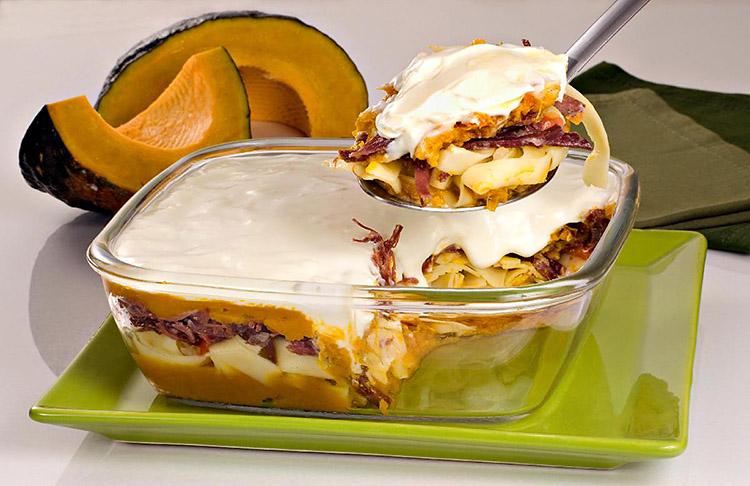 O talharim ao creme de abóbora com carne-seca está dentro de um refratário de vidro quadrado. É possível ver as camadas do prato e uma porção está sendo tirada com uma colher