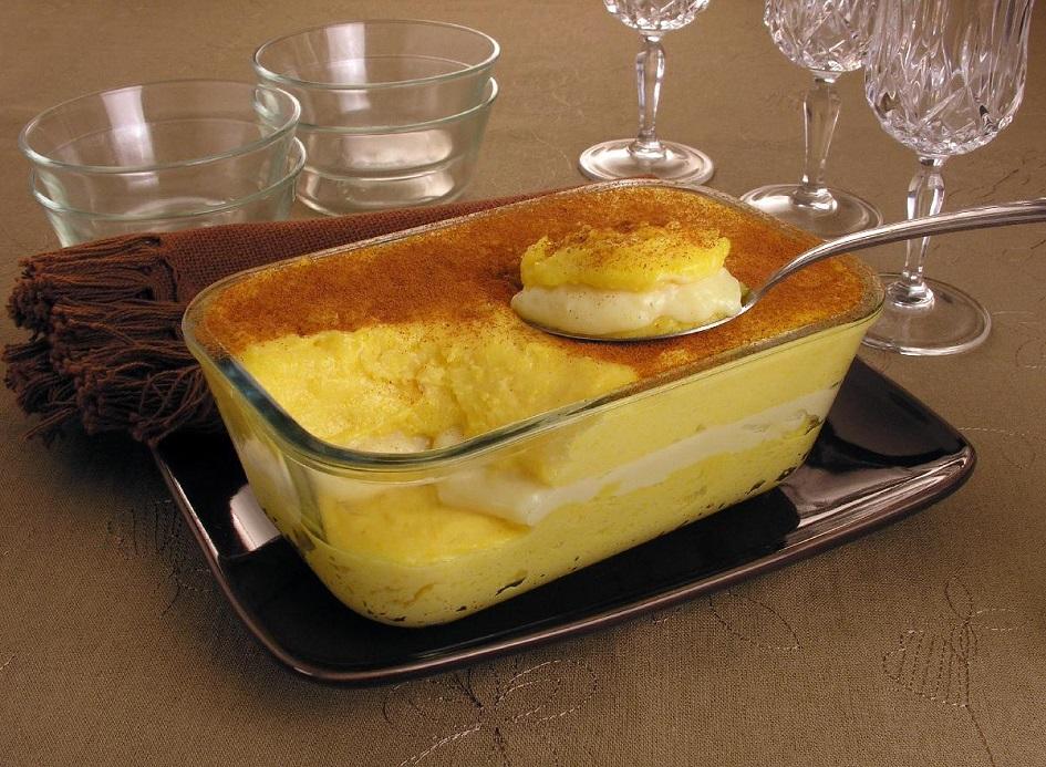 O curau recheado está dentro de uma travessa retangular de vidro e é possível ver as camadas e o recheio. O curau está polvilhado com canela e uma colher está tirando uma porção da sobremesa.