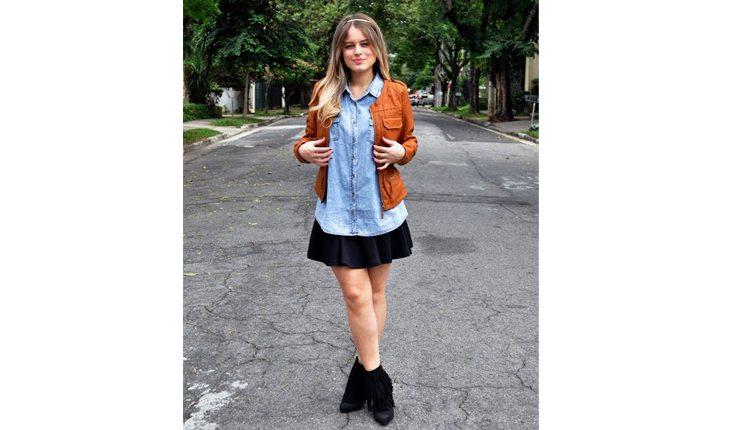 mulher com bota de cano curto preta, saia preta, camisa jeans e jaqueta marrom por cima