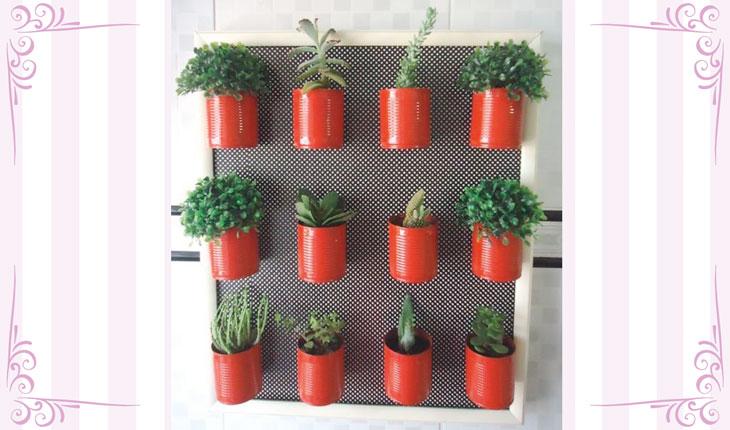 Na imagem há uma foto centralizada de um jardim vertical feito com latas que estão pintadas de vermelho. Em cada uma das latas há uma planta e o fundo é preto. Ao redor há uma moldura branca com detalhes em rosa.