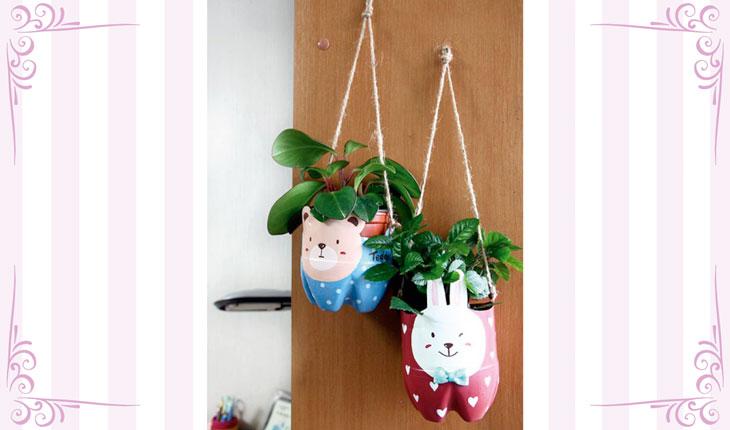 Na imagem há uma foto centralizada de dois vasinhos feitos com garrafa pet e pintados e decorados. Os vasinhos estão com plantas e pendurados verticalmente com uma corda. Ao redor há uma moldura branca com detalhes em rosa.