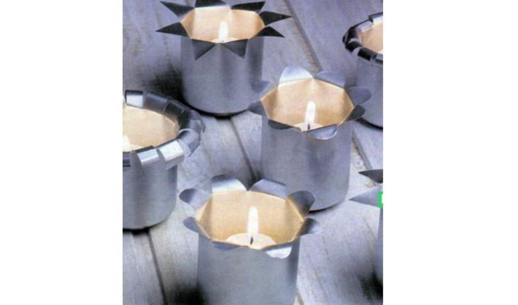 Na foto há vários suportes para vela feitos com latas de alumínio. Elas estão sem o desenho externo e são cortadas pela metade, com a parte do corte virada para fora para decorar. Há velas acesas dentro.