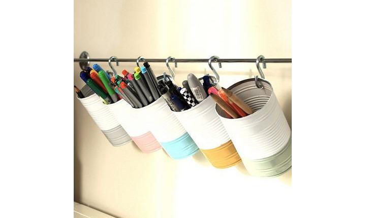 Na foto h´possível ver várias latas de aço pintadas metade de branco e metade (do fundo) de colorido. Elas estão penduradas em um suporte com ganchos e servem para armazenar canetas, lápis e outros materiais de escritório.