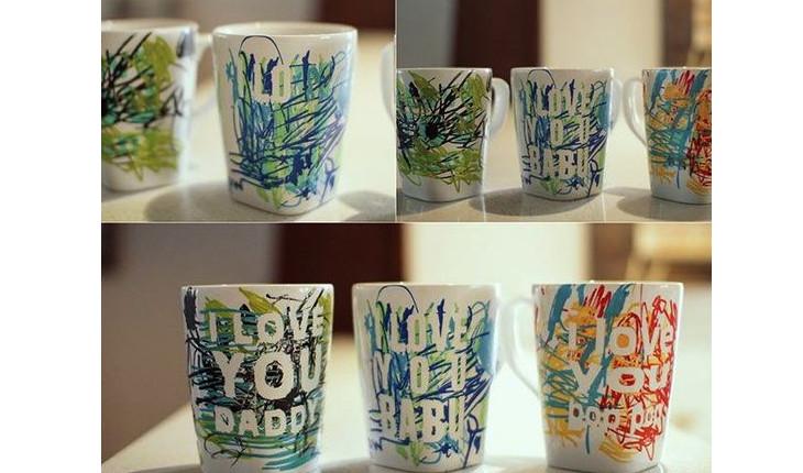 Na foto há duas imagens de canecas brancas decoradas com rabiscos coloridos feitos por crianças. Depois é tirado o adesivo que forma a frase e os locais onde estão as letras ficam em branco, formando a frase 