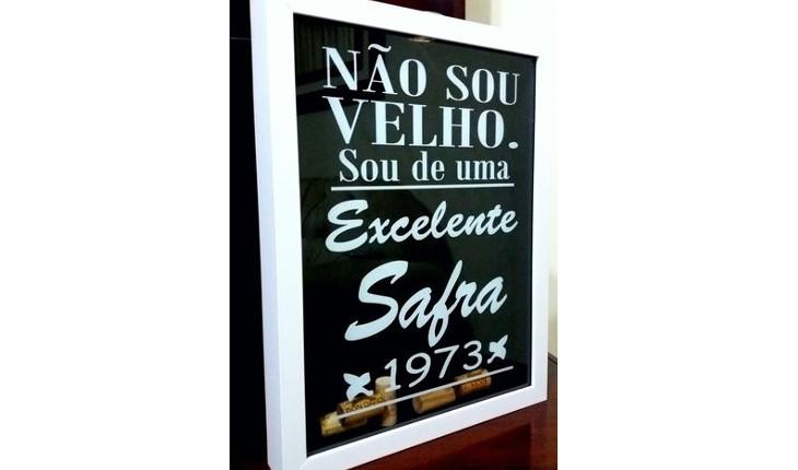 Na foto há uma quadro feito para armazenar rolhas de garrafas de vinho. A parte da frente do quadro é de vidro e está colada a frase 