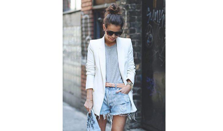 Na foto há uma mulher com shorts jeans largo, uma blusa cinza e um blazer branco.