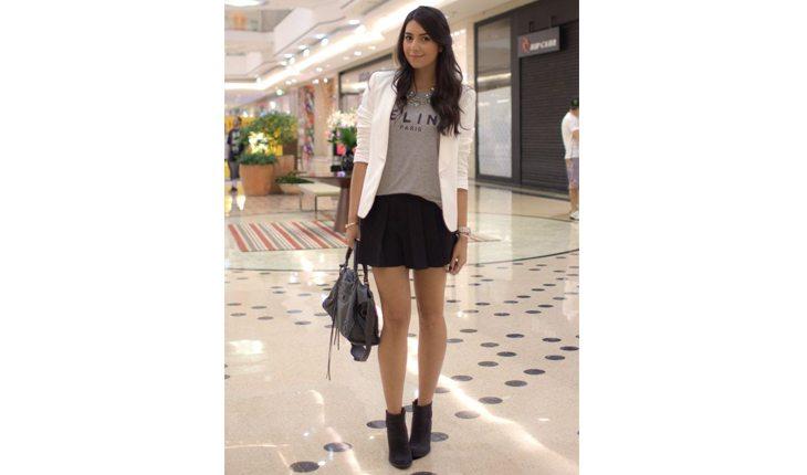 Na foto há uma mulher com saia preta, bota preta de cano curto, uma blusa cinza e um blazer branco por cima.