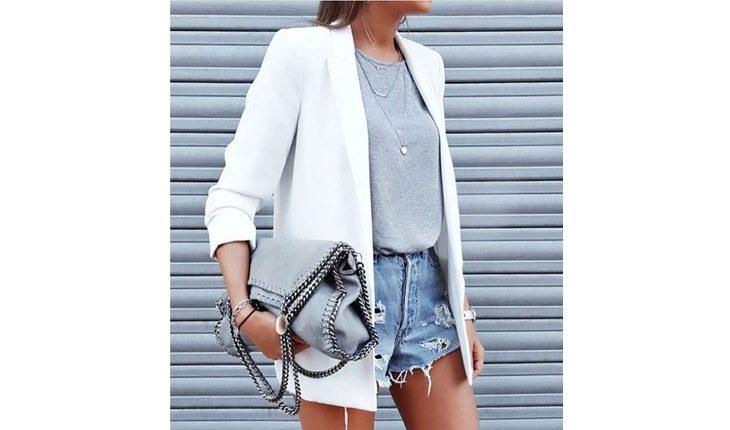Na foto há uma mulher com shorts jeans, uma blusa cinza e um blazer branco comprido.