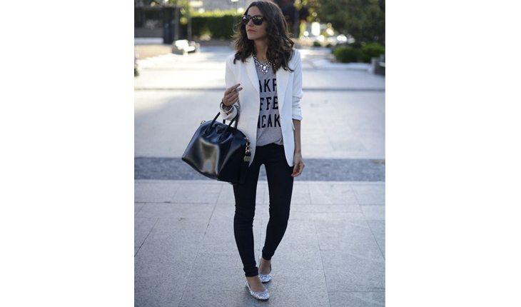 Na foto há uma mulher de calça preta, camiseta e blazer branco com uma bolsa preta.