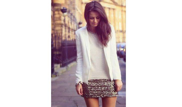 Na foto há uma mulher com uma saia de paetês prateada, uma blusa branca e um blazer branco.