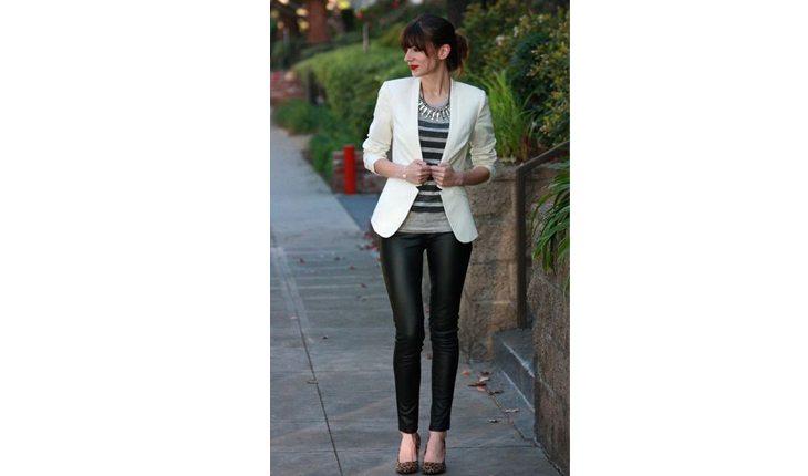 Na foto há uma mulher com calça preta, blusa cinza e um blazer branco. O sapato é preto e tem salto. O cabelo está em coque.