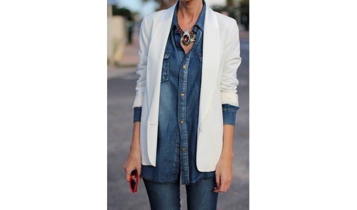 Na foto há uma mulher com camisa jeans e um blazer branco por cima dobrado no meio do braço.