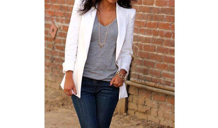 Na foto há uma mulher de calça jeans, uma blusa cinza e um blazer branco. Ela está com um colar fino e comprido.