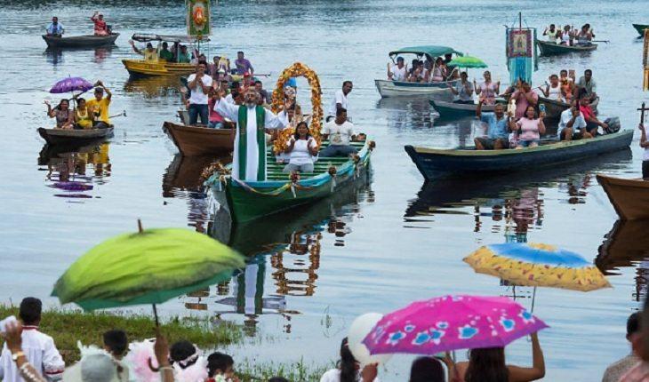 Vários barcos estão no rio, em um, a santa vai atrás de um padre. Nossa Senhora de Nazaré.